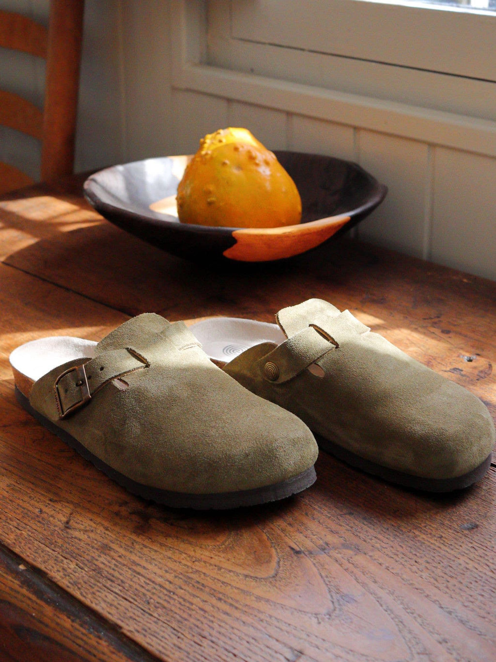 GENUINS RIVA Velour Clogs Khaki – Lifestyle-Shot auf Holztisch mit Obstschale, warme Herbststimmung